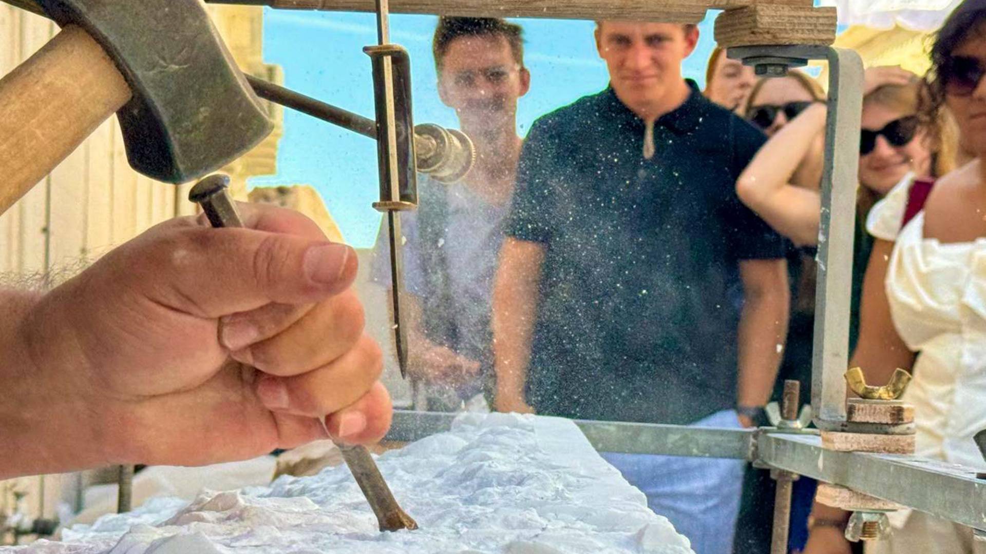 A person's hand chiseling a marble block, a group of students observing carefully.