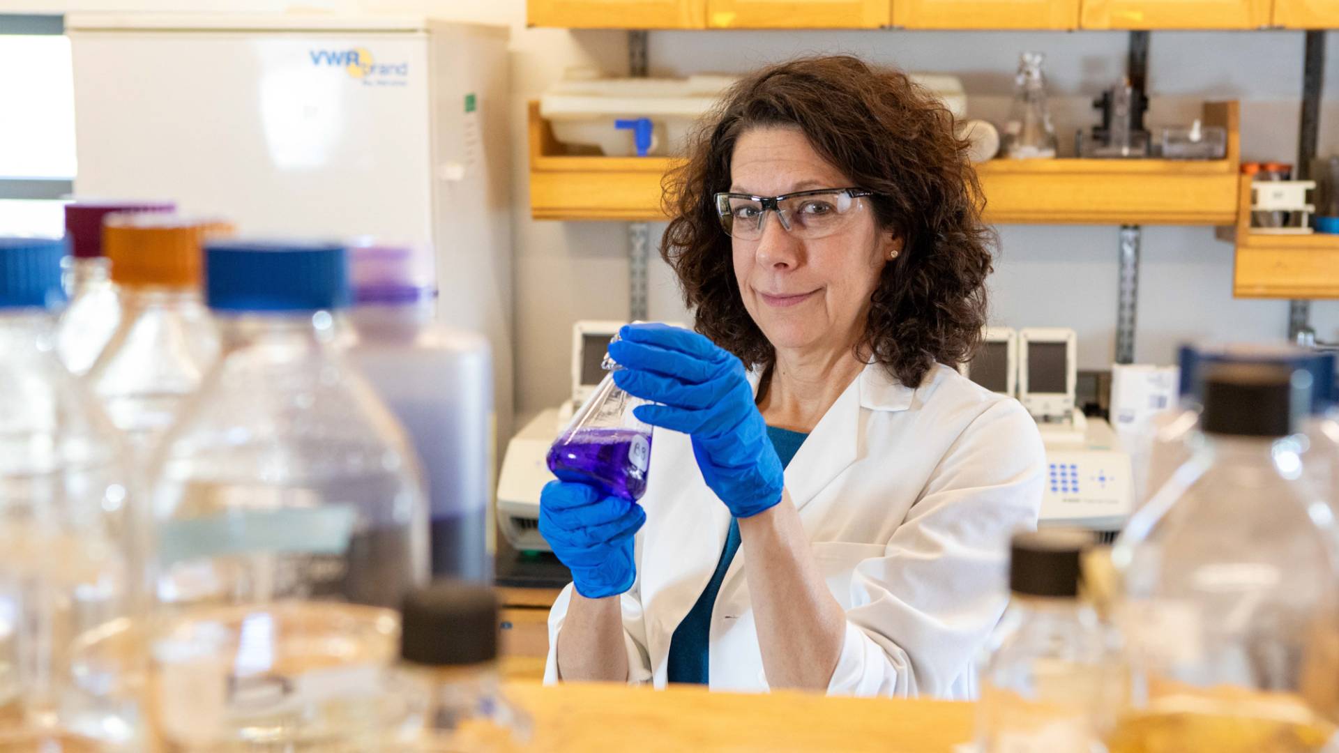 Bonnie holding in a lab wearing eye protective wear, and gloves holding an apparatus.