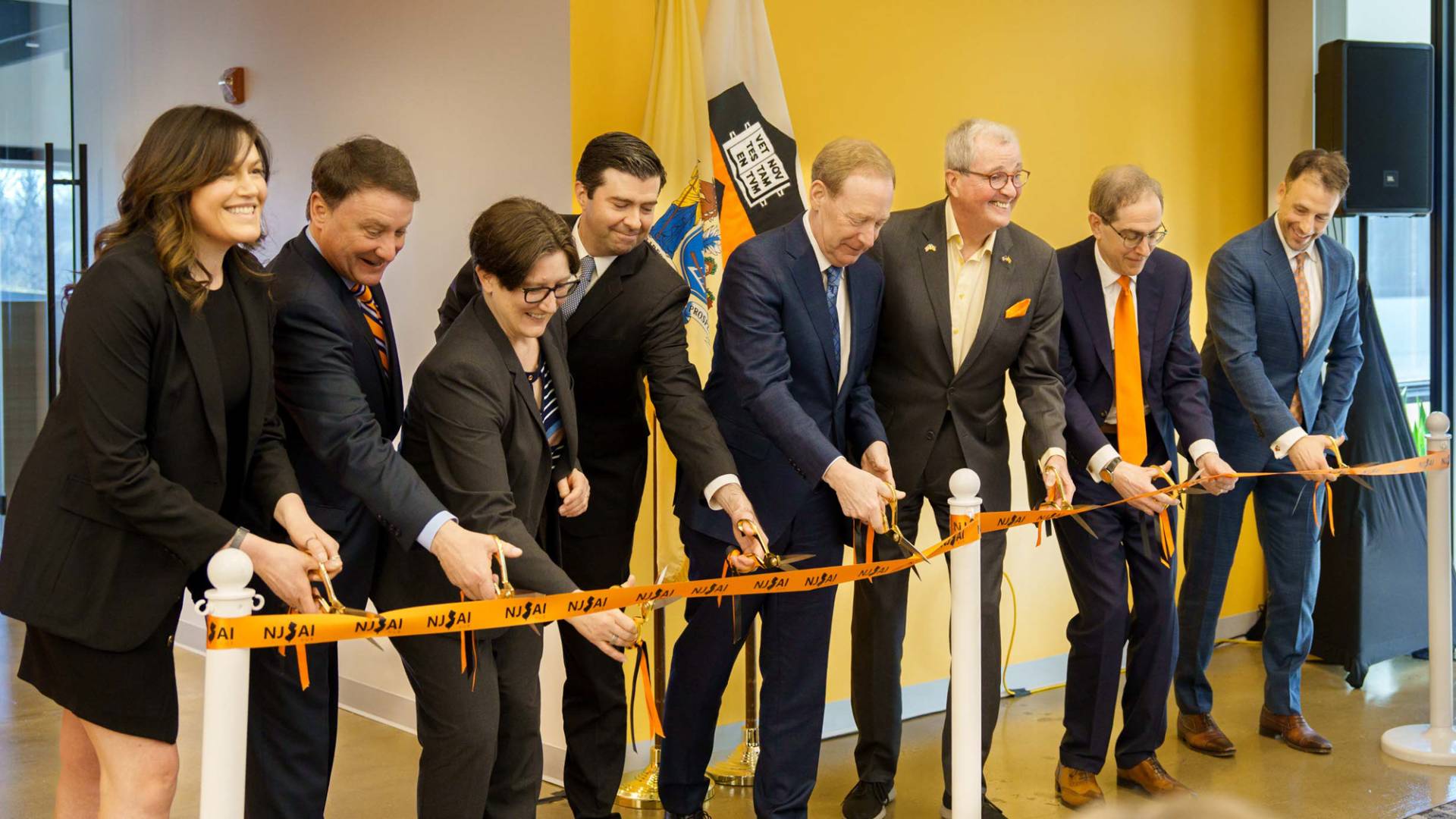 A line of smiling men and woman hold scissors up to a bright orange ribbon.