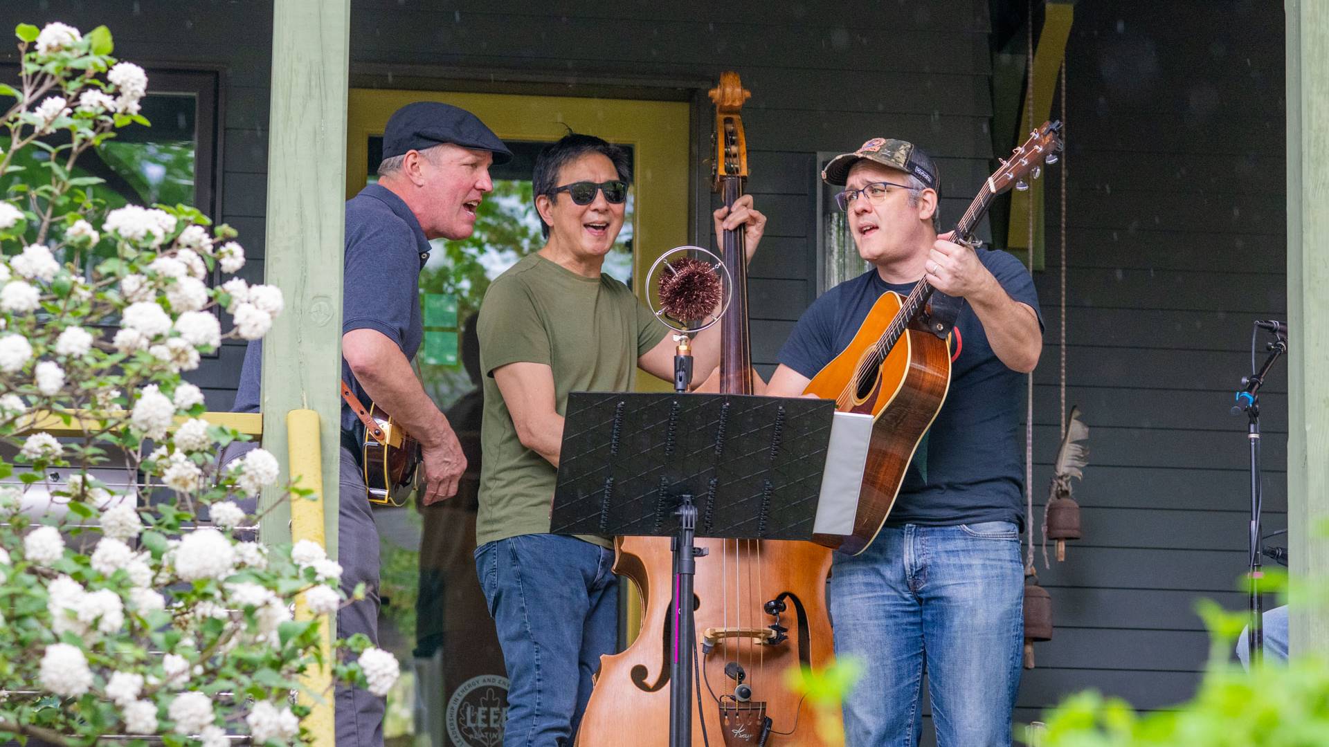 A trio of performers playing banjo, upright bass and guitar read sheet music from a stand outside a blue-green residential porch.