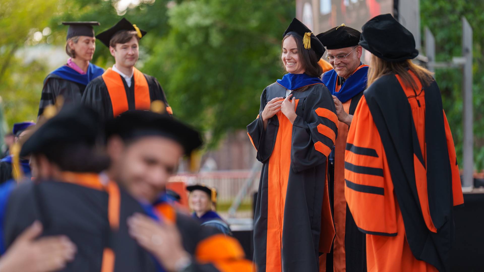 Three sets of graduates and their advisors -- one hugging in the foreground, a second set actively hooding in the center, and a third pair waiting on the side.