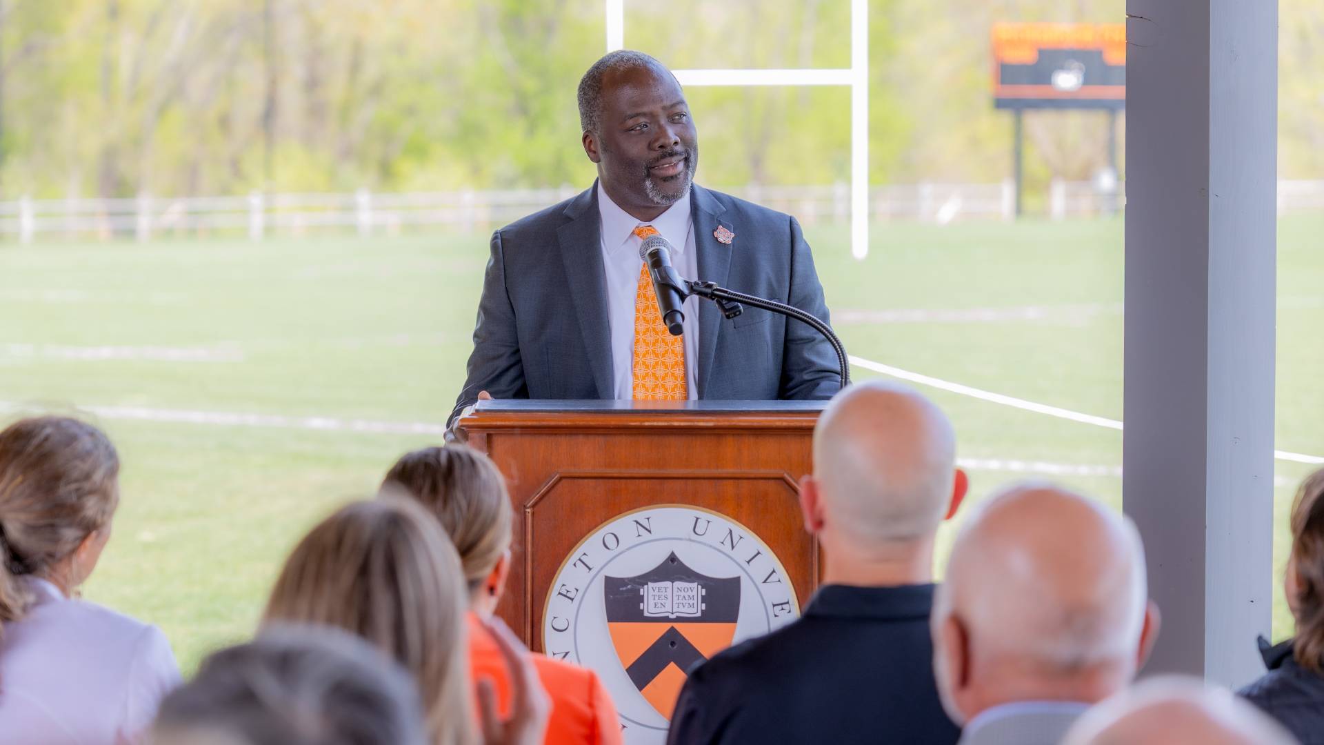John Mack speaks at podium with Rickerson Field in the background during the ribbon-cutting for the rugby facilities at Haaga House.