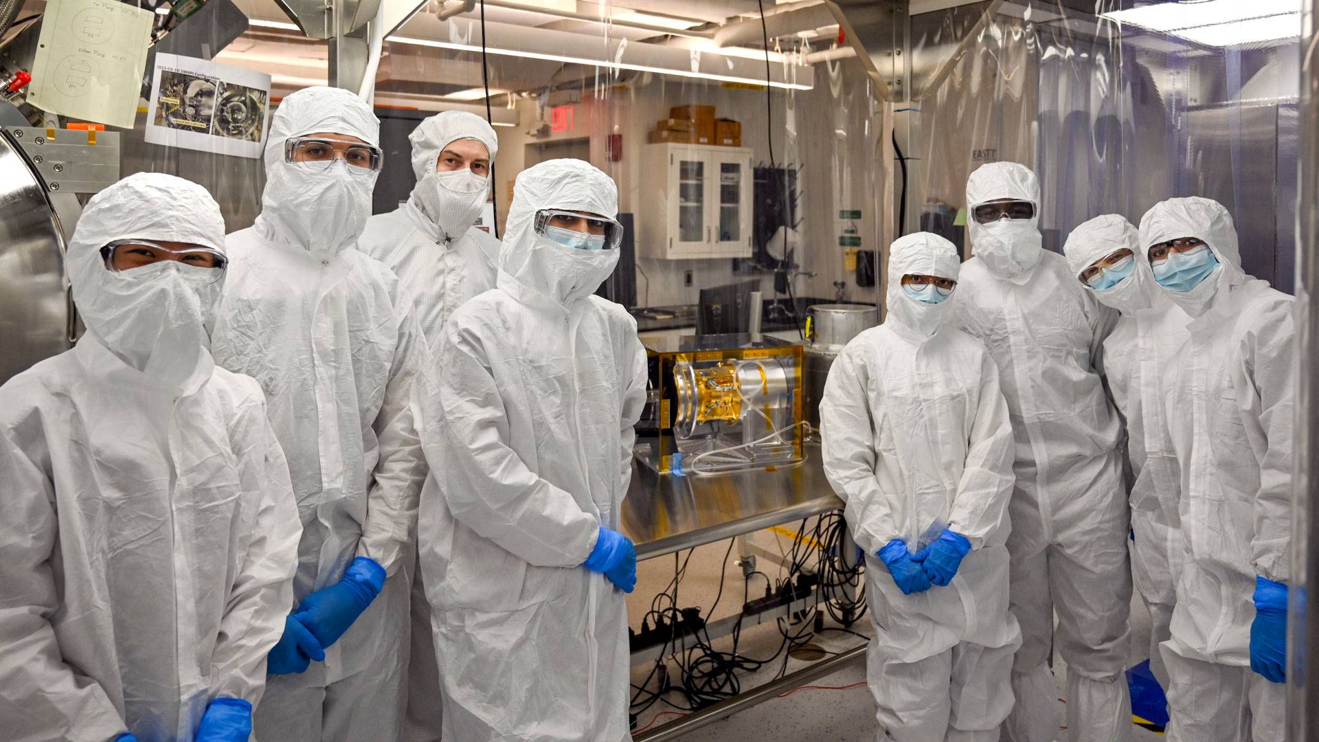 A group of students and technicians in white coveralls stand around a small golden instrument.