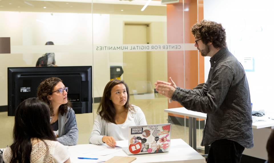 A photo of a teacher speaking to a small group of students seated at a table.