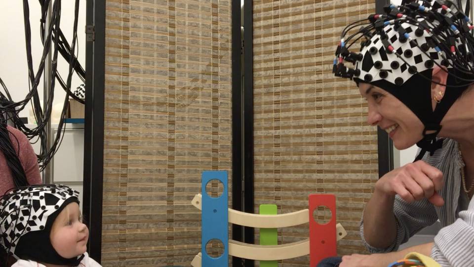 A baby and a researcher wear brain wavelength-measuring devises on their heads while making eyecontact during play