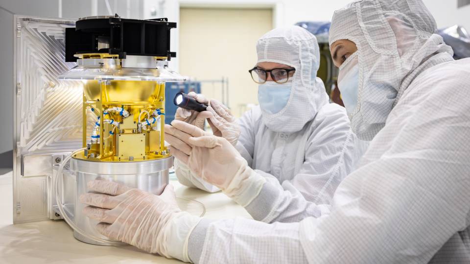 Scientists in cleanroom gear look at a golden instrument