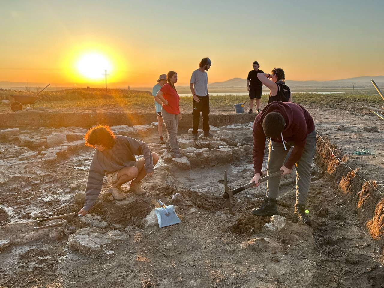 A group of people digging in the dirt, sunset behind them.