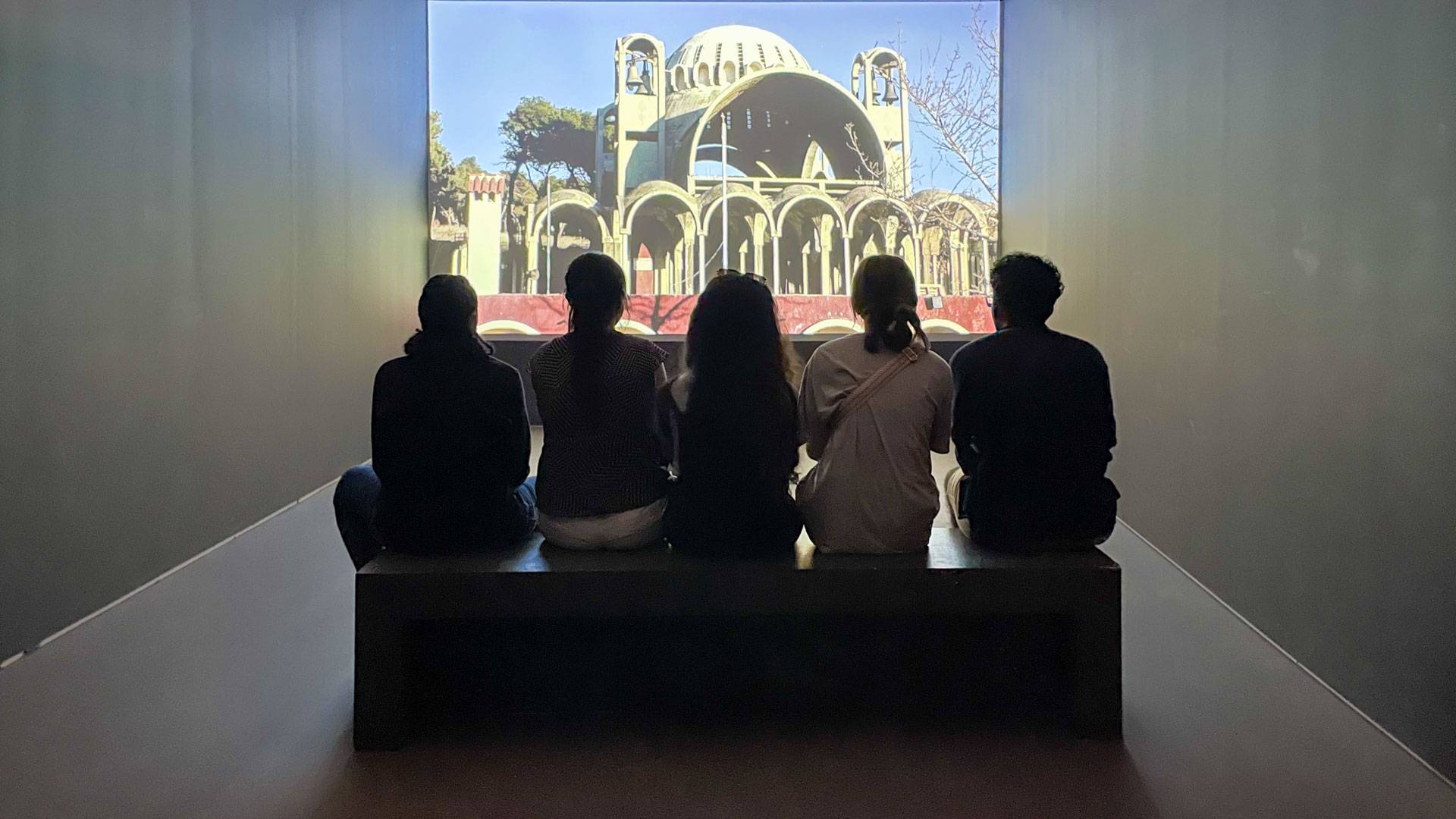 The backs of four people seated in front of an art museum display
