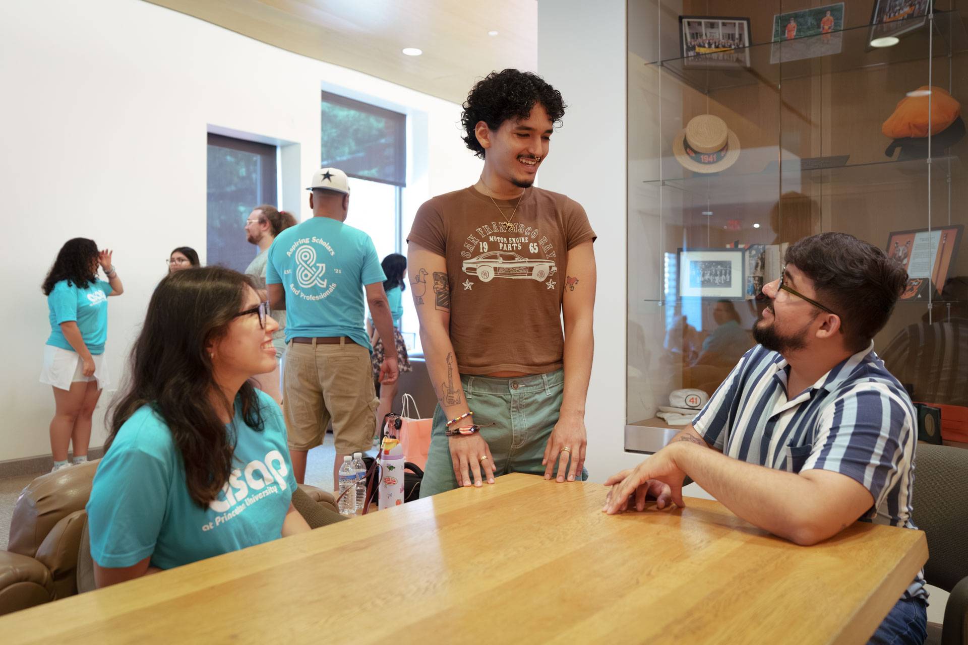 Student interns, one sitting and the other standing, engage in conversation with their mentor at a wood table