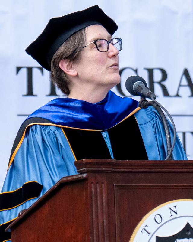 A woman in a blue doctoral robe speaks at a podium.