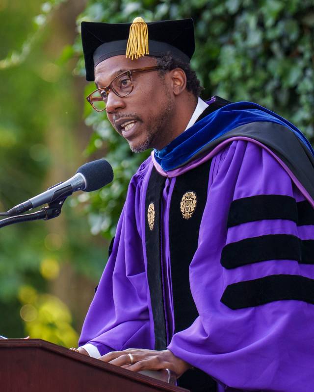 A man in purple doctoral robes speaks into a microphone.